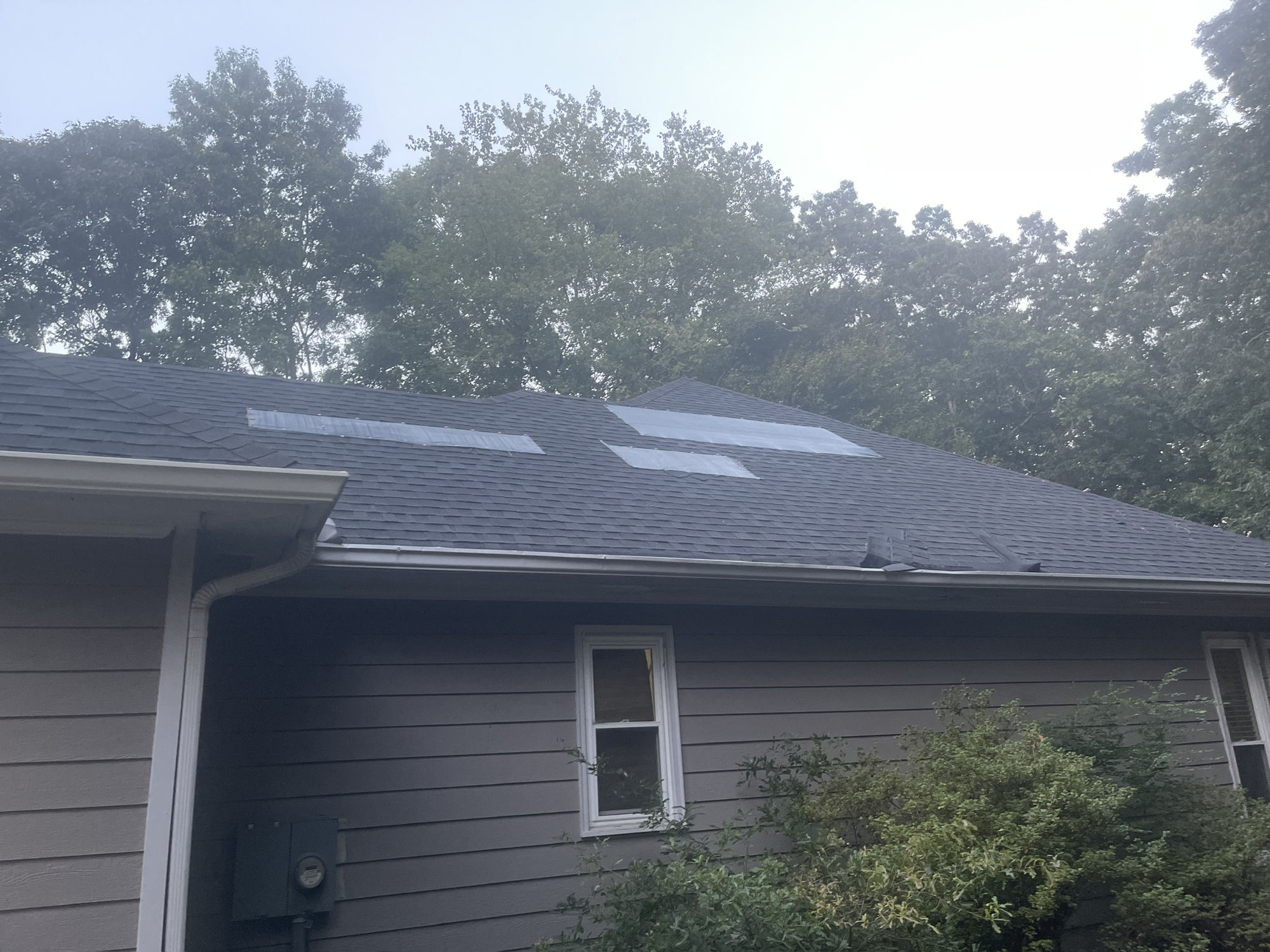 Rear slope. Several membrane patch sections mid-slope and torn shingle material hanging over the gutter line at the right eave.